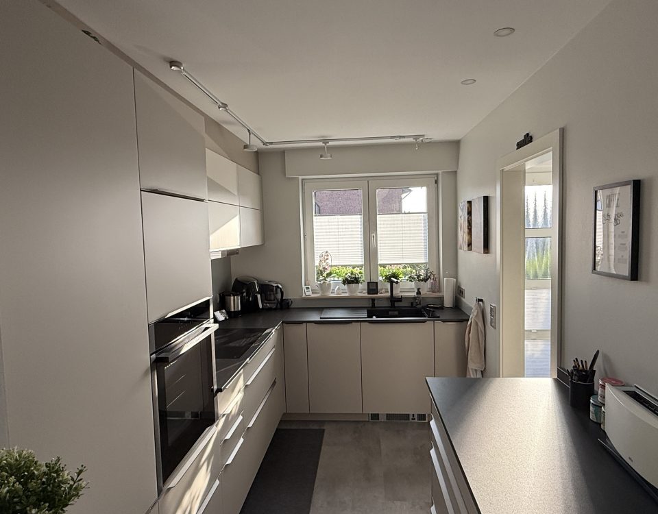 U-Küche mit SMARTGLAS-Fronten in Sandgrau, dunkler Neolith-Arbeitsplatte und Fensterzeile.