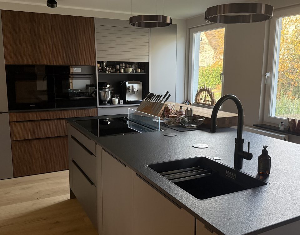 Kücheninsel mit dunkler Keramikplatte, aufgesetzter Eiche-Bar, schwarzer Spüle und Armatur, im Hintergrund Schrankzeile.