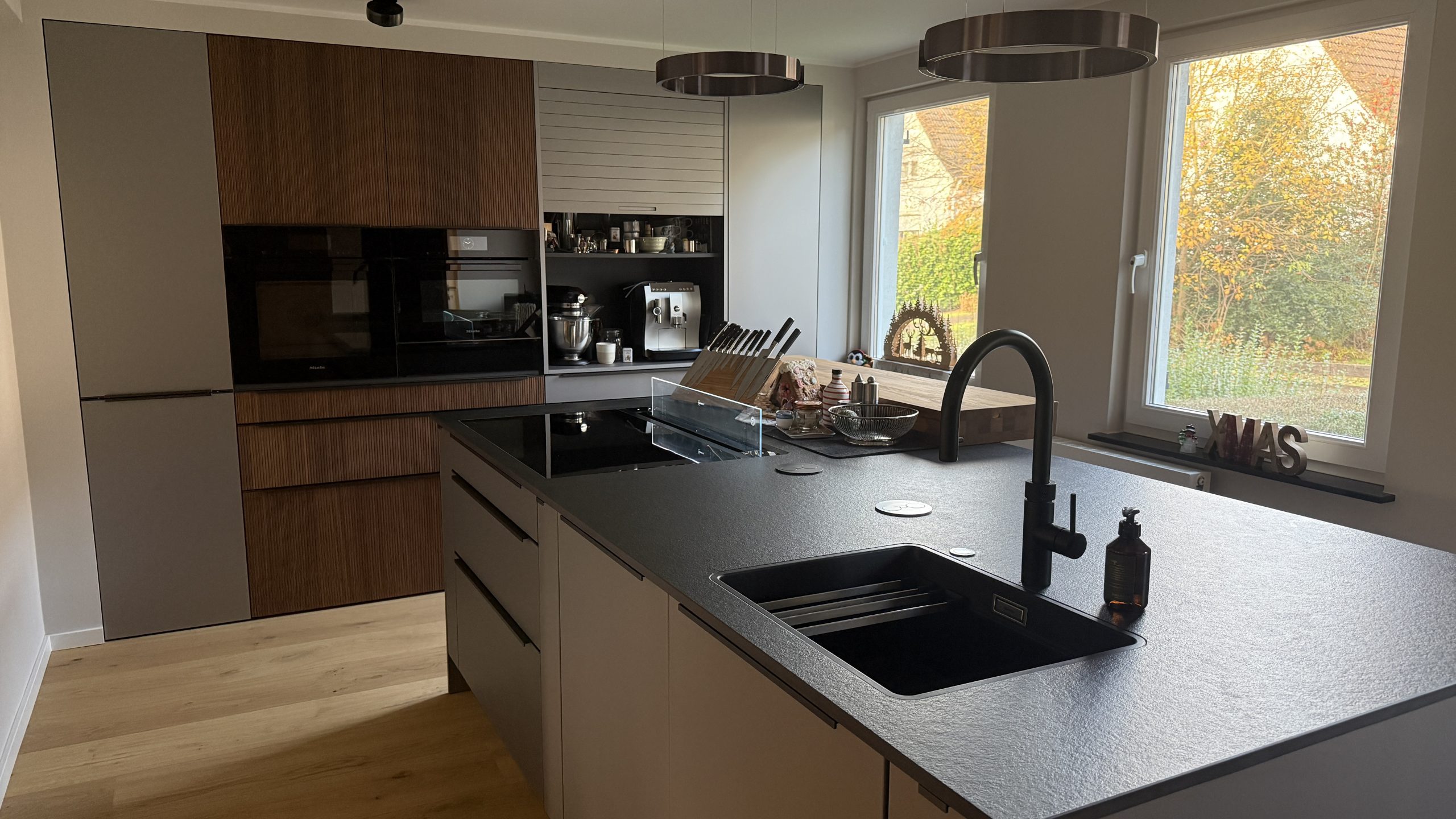 Kücheninsel mit dunkler Keramikplatte, aufgesetzter Eiche-Bar, schwarzer Spüle und Armatur, im Hintergrund Schrankzeile.