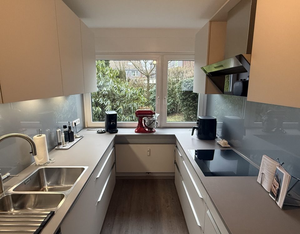 Küchenzeile mit satinierten Glasfronten in Weiß, grauer Quarzkomposit-Arbeitsplatte und weißer Glasrückwand; Spüle, Induktion und Unterbauleuchten.