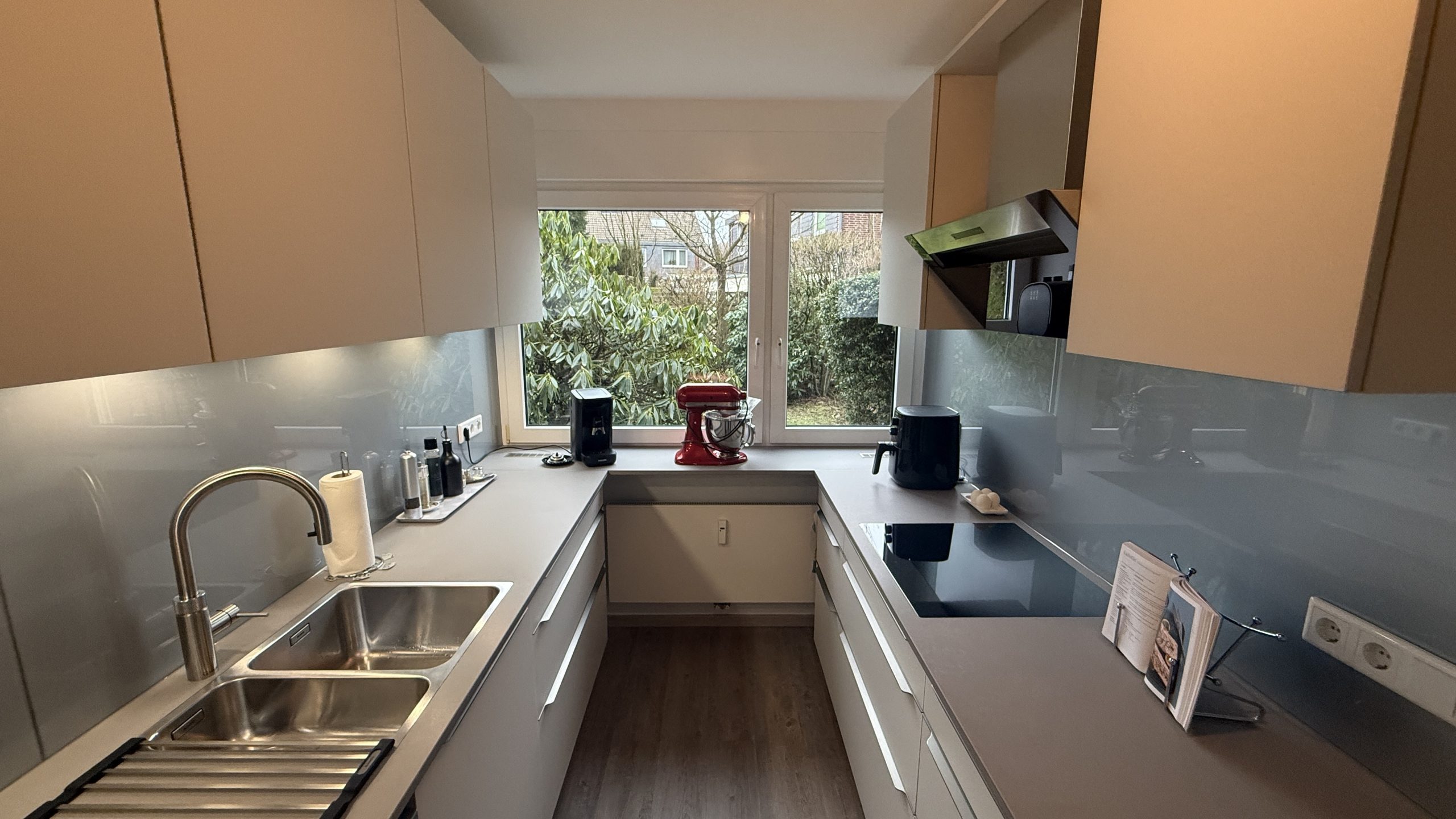 Küchenzeile mit satinierten Glasfronten in Weiß, grauer Quarzkomposit-Arbeitsplatte und weißer Glasrückwand; Spüle, Induktion und Unterbauleuchten.