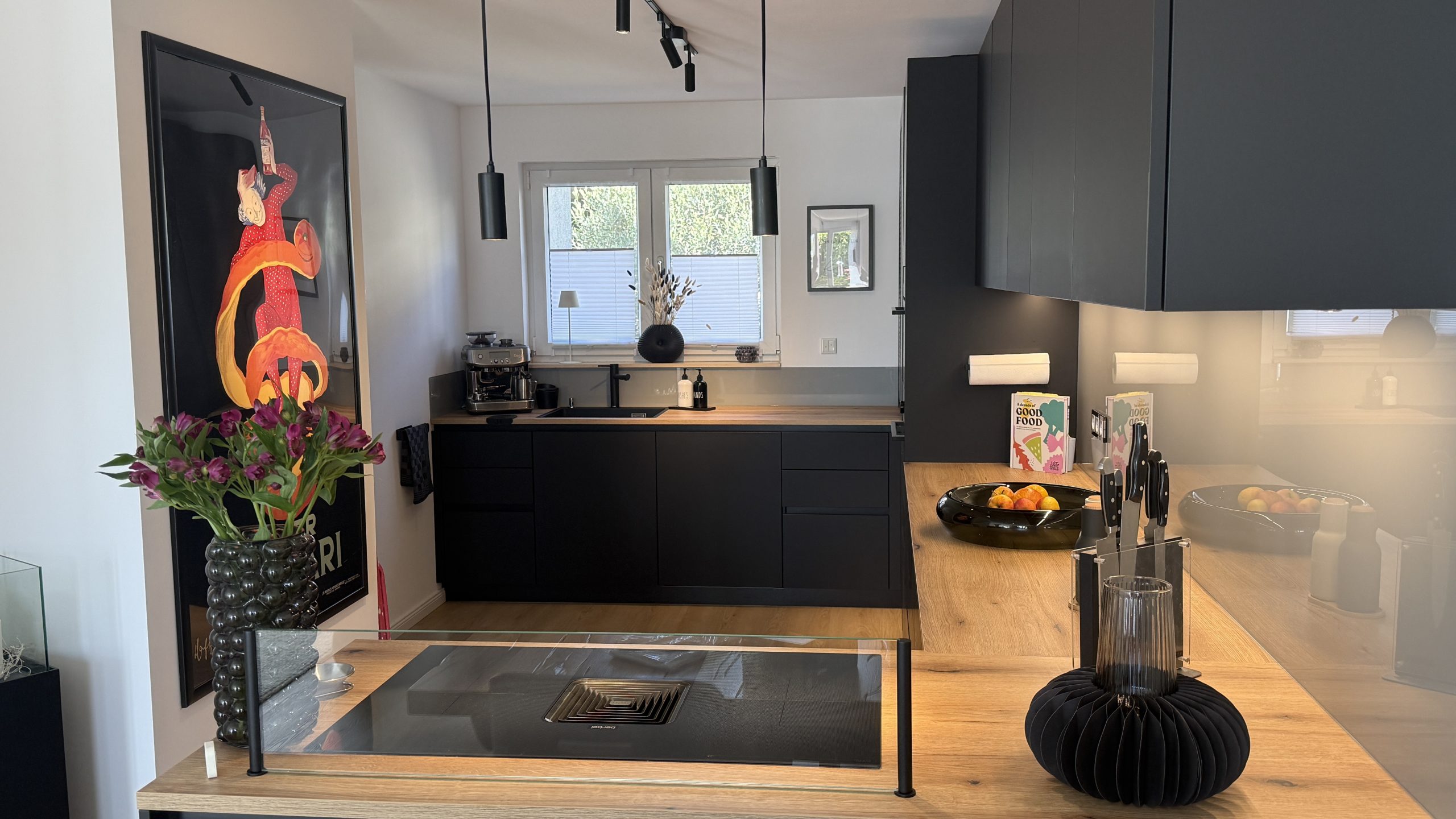 Schwarze Küche mit Holzarbeitsplatte, Kochfeld mit Glasaufsatz und Fensterzeile im Hintergrund.