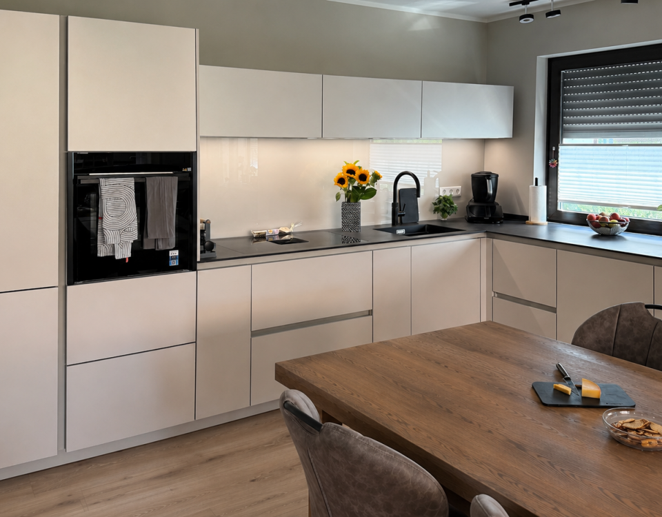 U-förmige Küche mit hellen Silk-Metallic-Fronten, schwarzer Dekton-Arbeitsplatte, schwarzer Spüle und Holztisch im Vordergrund.