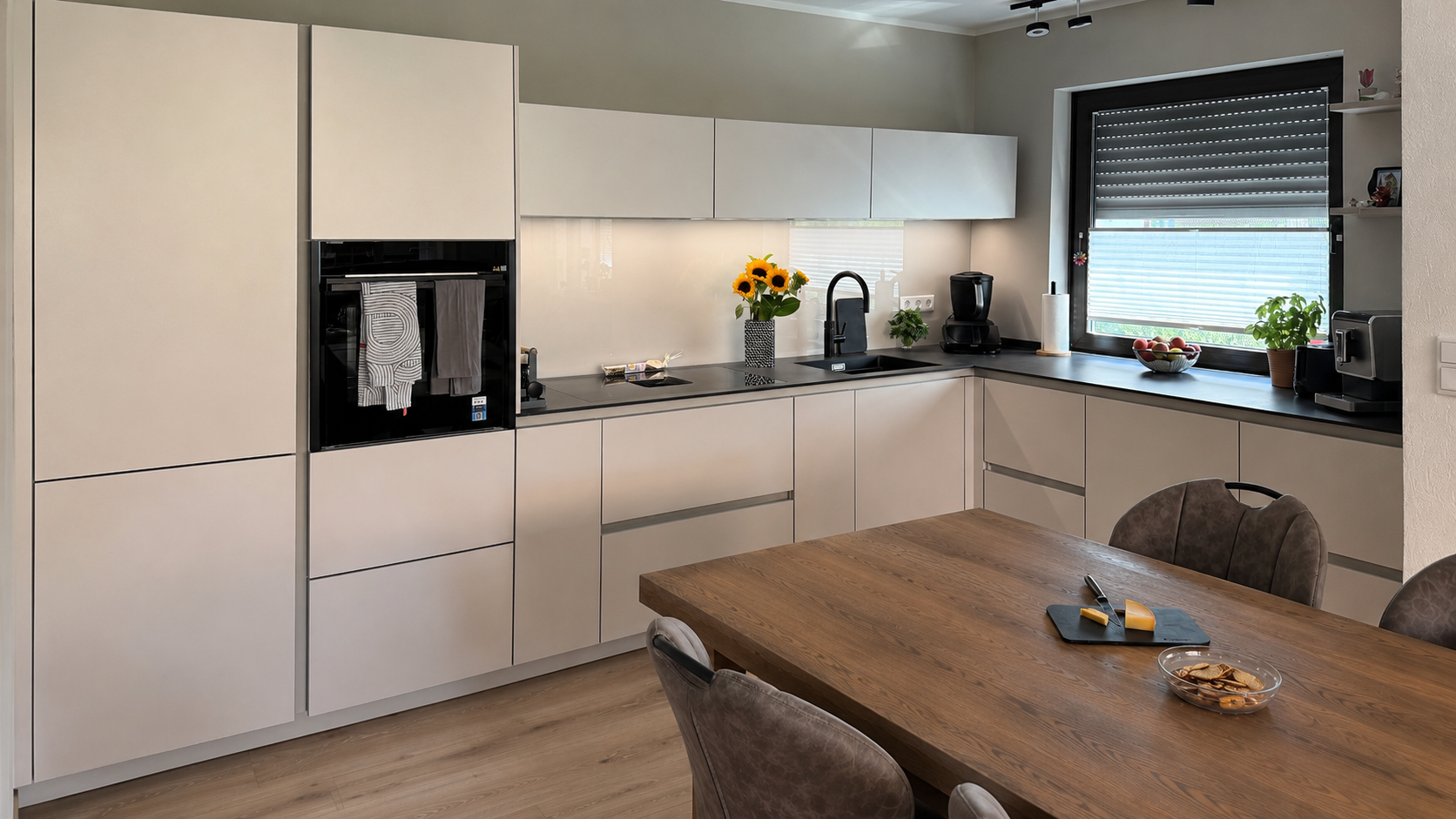 U-förmige Küche mit hellen Silk-Metallic-Fronten, schwarzer Dekton-Arbeitsplatte, schwarzer Spüle und Holztisch im Vordergrund.