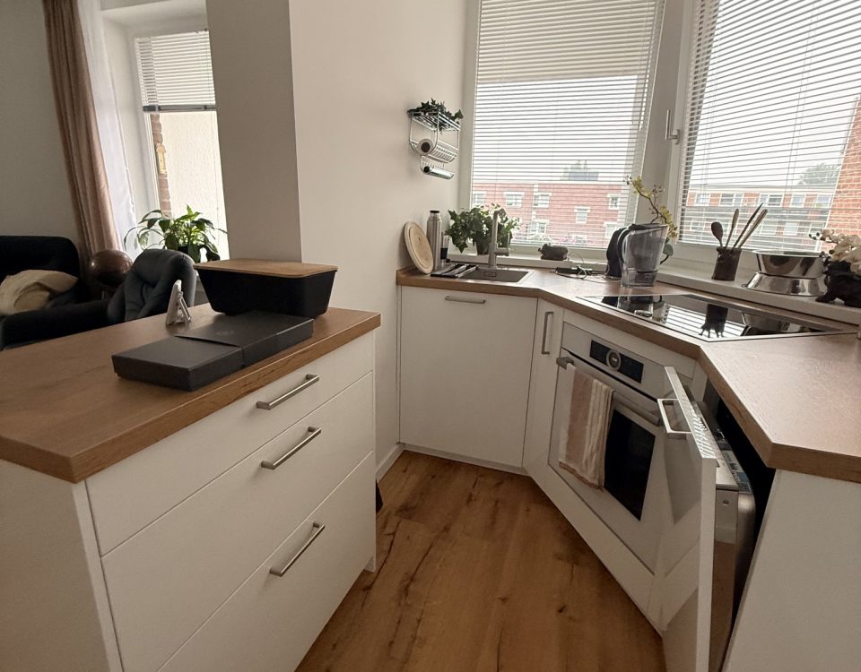 Blick zwischen zusätzlichem Arbeitsblock und weißer U-Küche mit Holzarbeitsplatte und großen Fenstern.