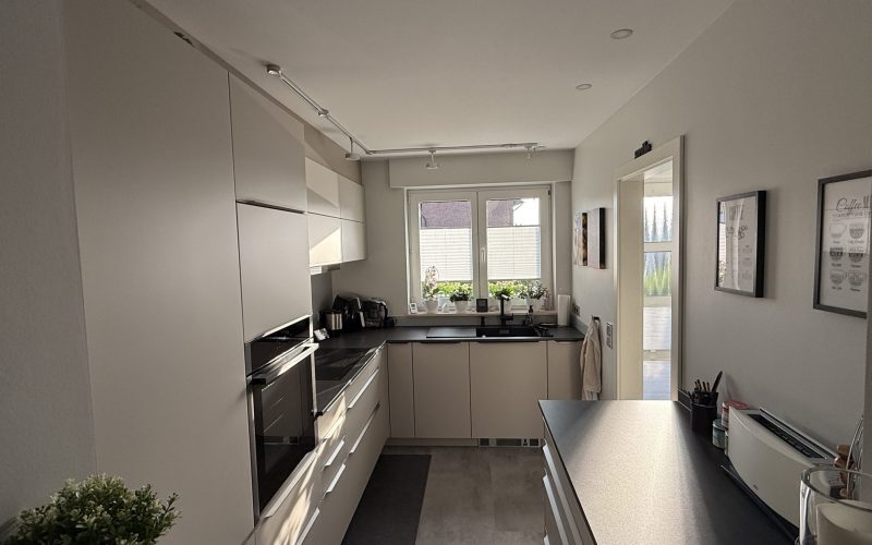 U-Küche mit SMARTGLAS-Fronten in Sandgrau, dunkler Neolith-Arbeitsplatte und Fensterzeile.
