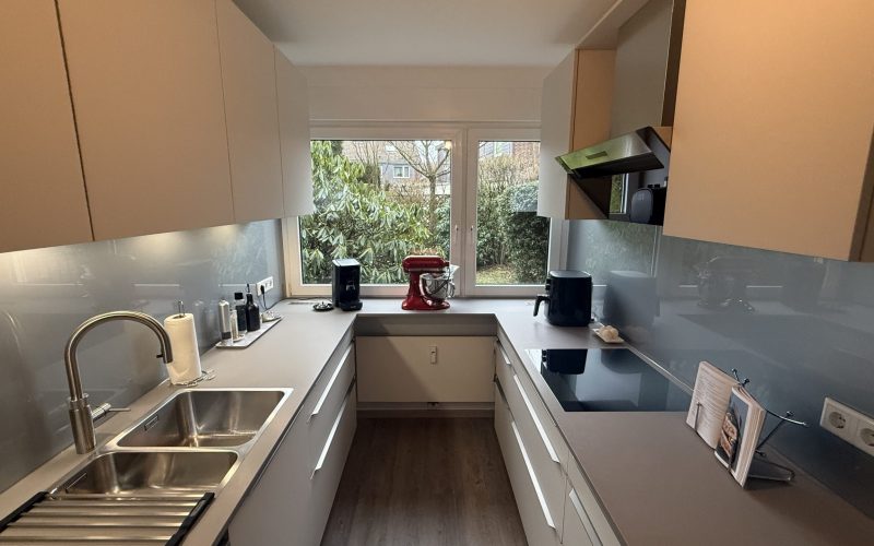 Küchenzeile mit satinierten Glasfronten in Weiß, grauer Quarzkomposit-Arbeitsplatte und weißer Glasrückwand; Spüle, Induktion und Unterbauleuchten.