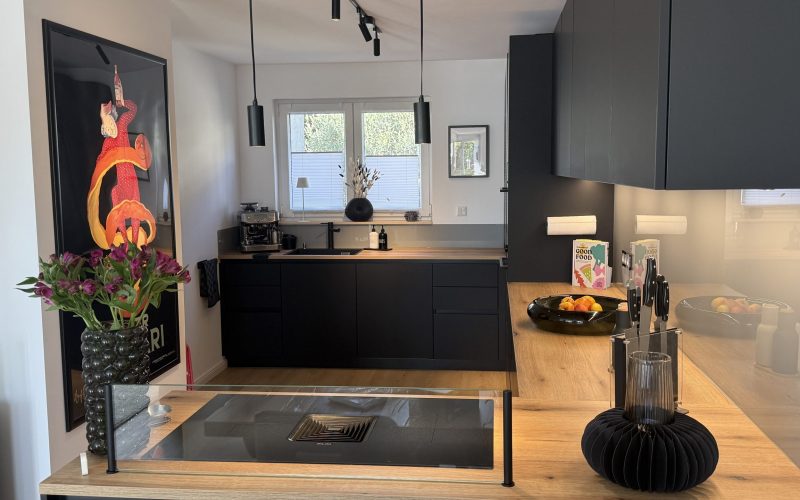 Schwarze Küche mit Holzarbeitsplatte, Kochfeld mit Glasaufsatz und Fensterzeile im Hintergrund.