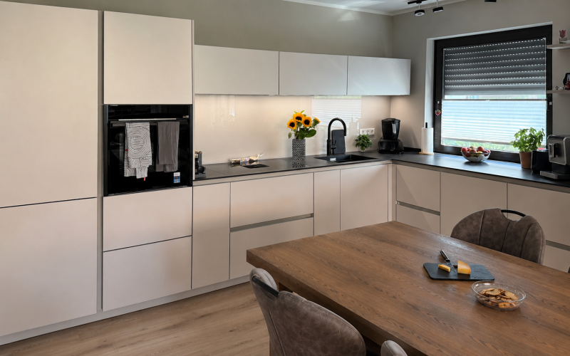 U-förmige Küche mit hellen Silk-Metallic-Fronten, schwarzer Dekton-Arbeitsplatte, schwarzer Spüle und Holztisch im Vordergrund.